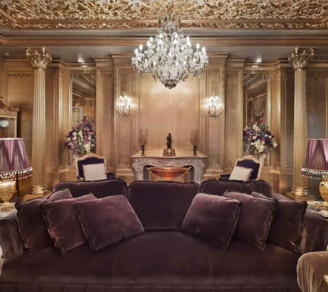 Salón lujoso con sofás de terciopelo morado, lámparas doradas y un candelabro de cristal. El techo y las paredes doradas evocan el estilo barroco español.