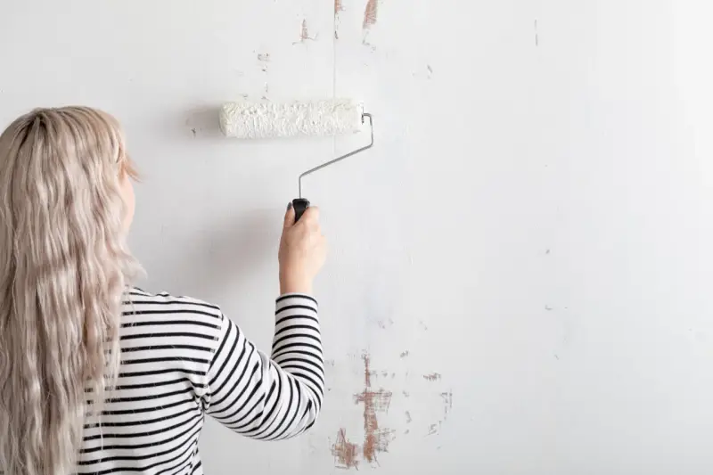 Mujer con cabello rubio y camiseta a rayas está a punto de empapelar pared con rodillo blanco.
