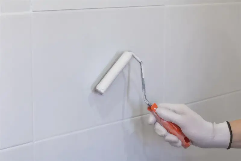 Mano enguantada pintando con rodillo blanco sobre azulejos grises. Renovando el aspecto de los tubos calefacción.