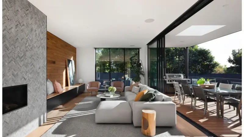 Elegante salón con chimenea de piedra, pared de madera y grandes ventanales que conectan con una terraza. Un ejemplo de las últimas tendencias interiorismo.