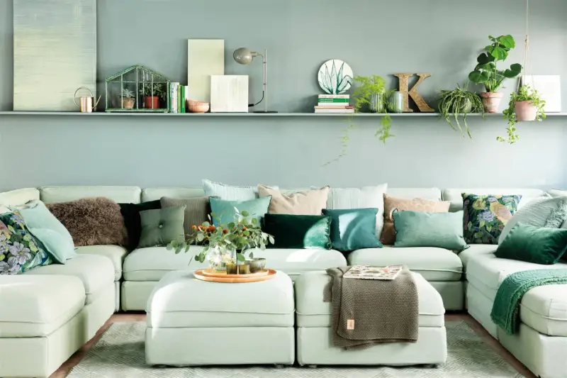 Sofá modular claro con cojines verdes y estampados, y una estantería con plantas y objetos decorativos, incluyendo una casita de cristal con **muebles pintados en verde**.